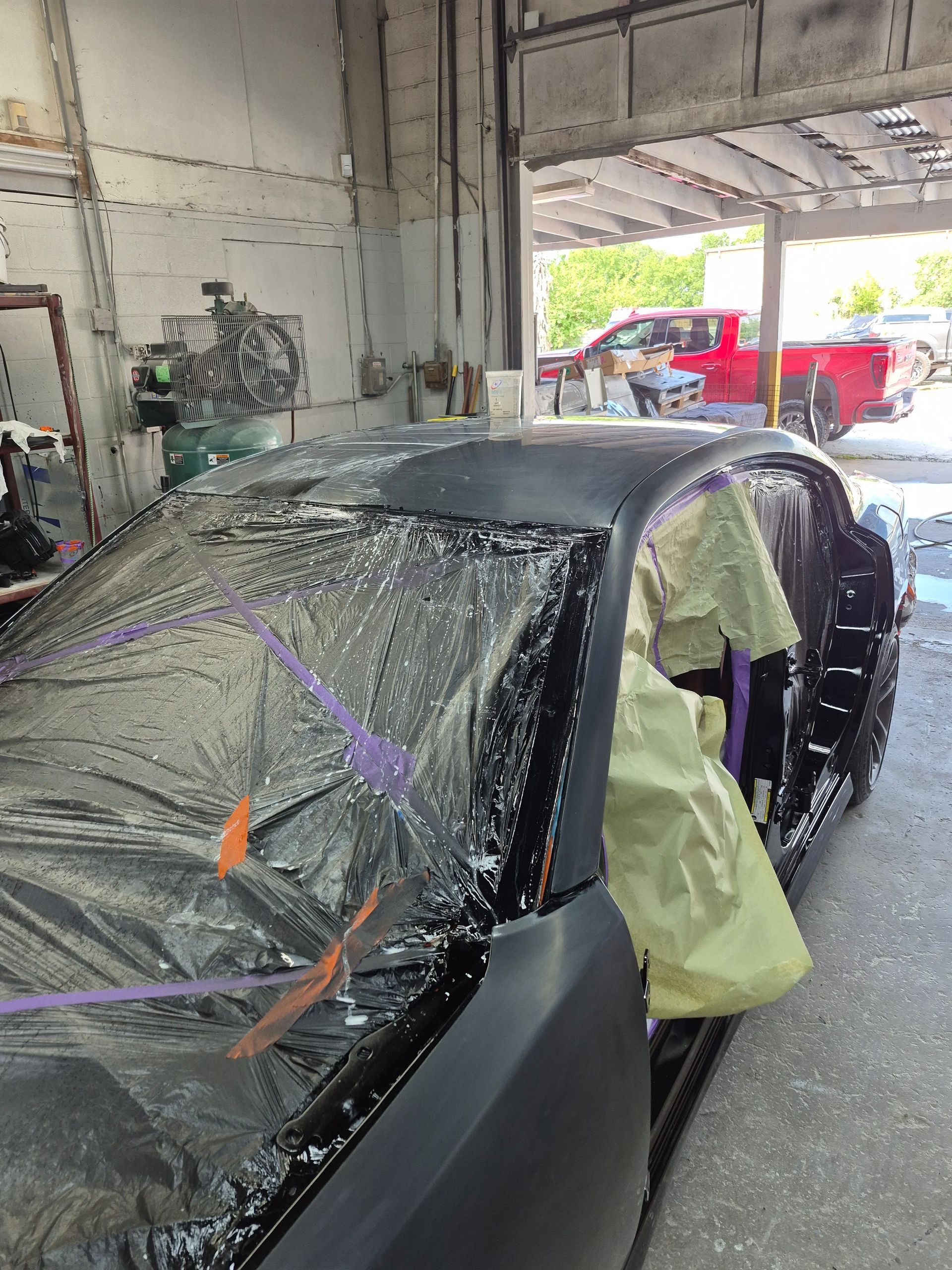 Car wrapped in plastic and tape inside a paint shop, with another red truck outside.