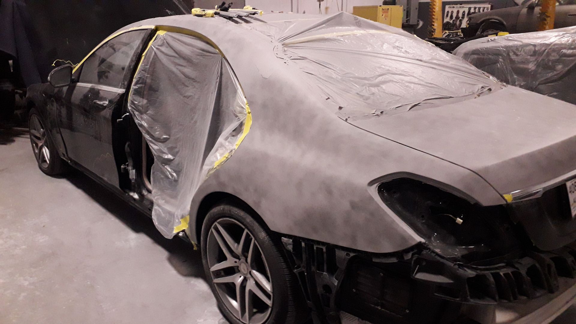 Gray sedan covered in dust and masking plastic in a body shop, with a door open and front bumper removed