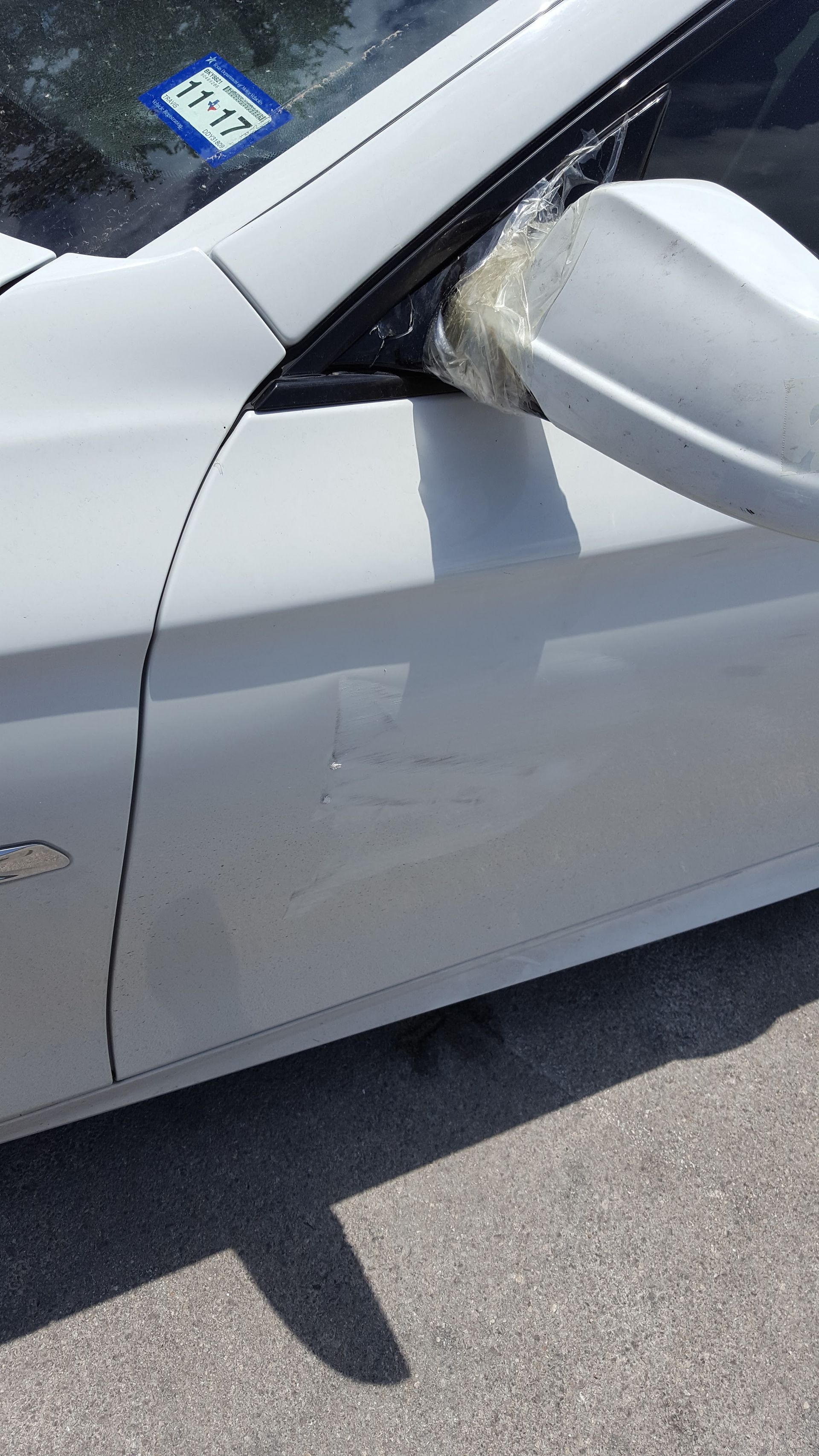 Close-up of a white car’s front side with a side mirror and visible body damage near the door seam.