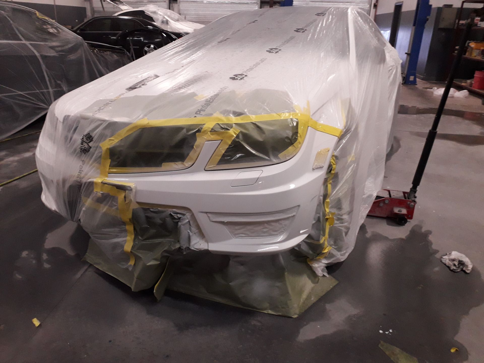 White car front covered in plastic and masking tape in a body shop, with bumper repair in progress