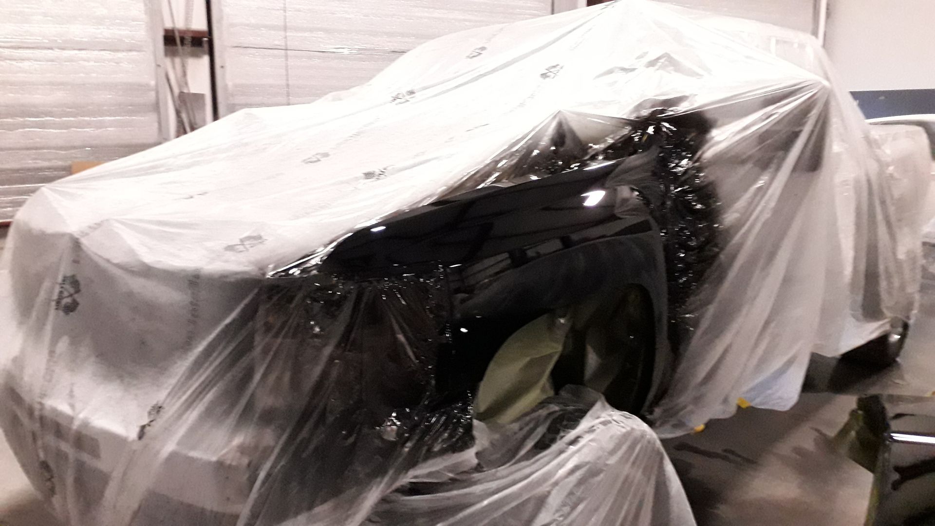 Car wrapped in white plastic sheeting inside a garage or workshop, with exposed front end.