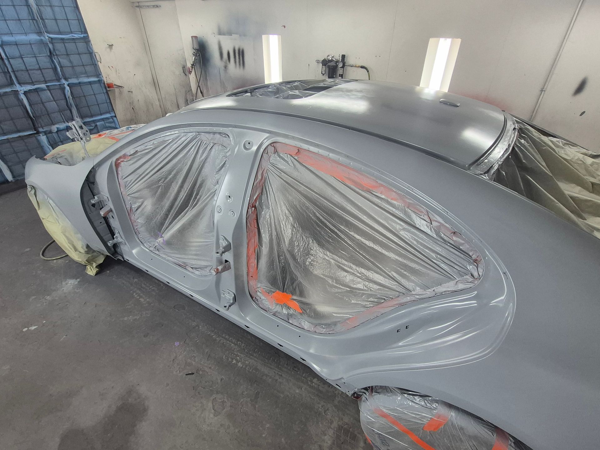 Gray sedan masked for paint in an auto body shop, with windows and trim covered in plastic and tape