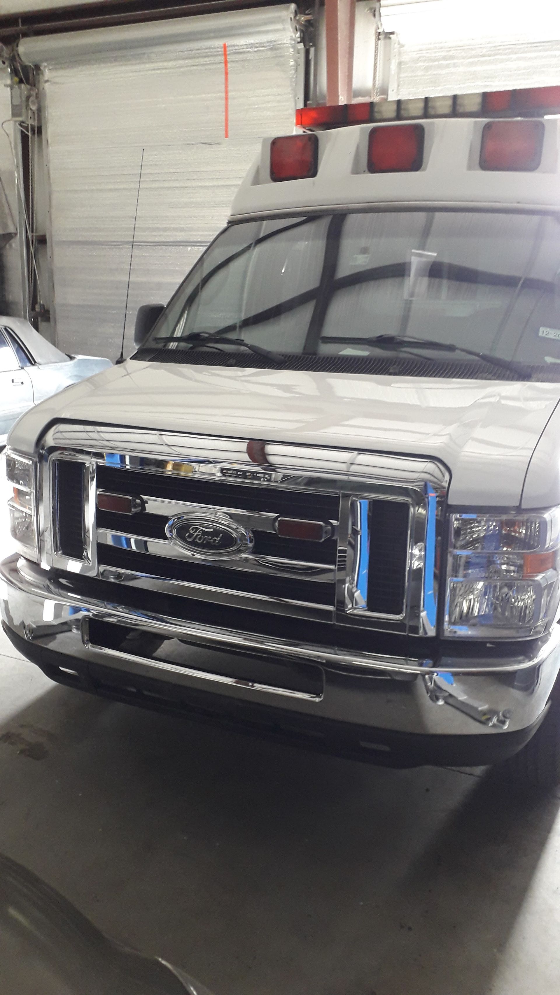 White emergency vehicle parked indoors, front view with red lights on the roof and blue-striped grille.
