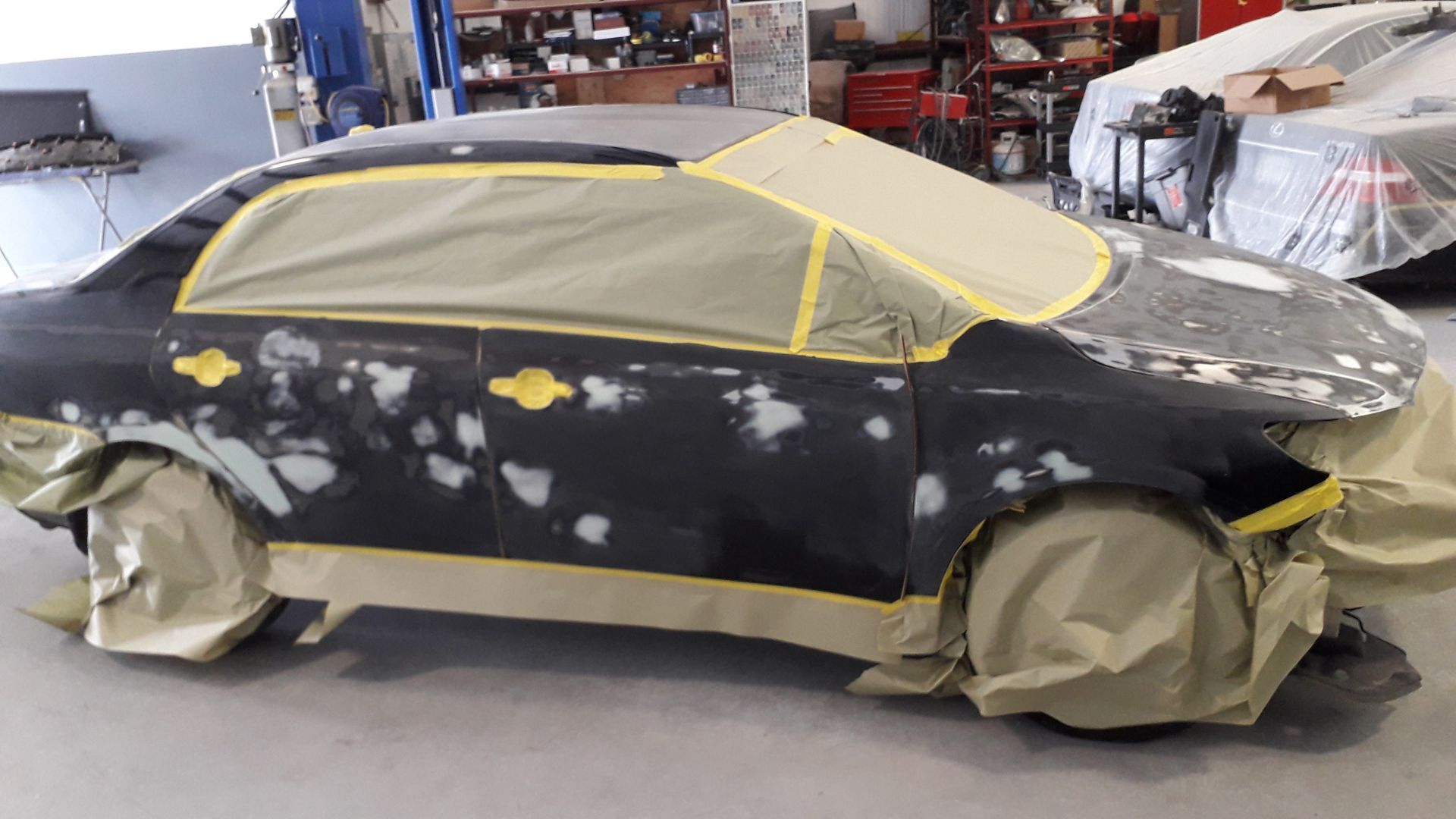 Black car in auto shop, covered in primer patches and masking tape, with windows and wheels taped off