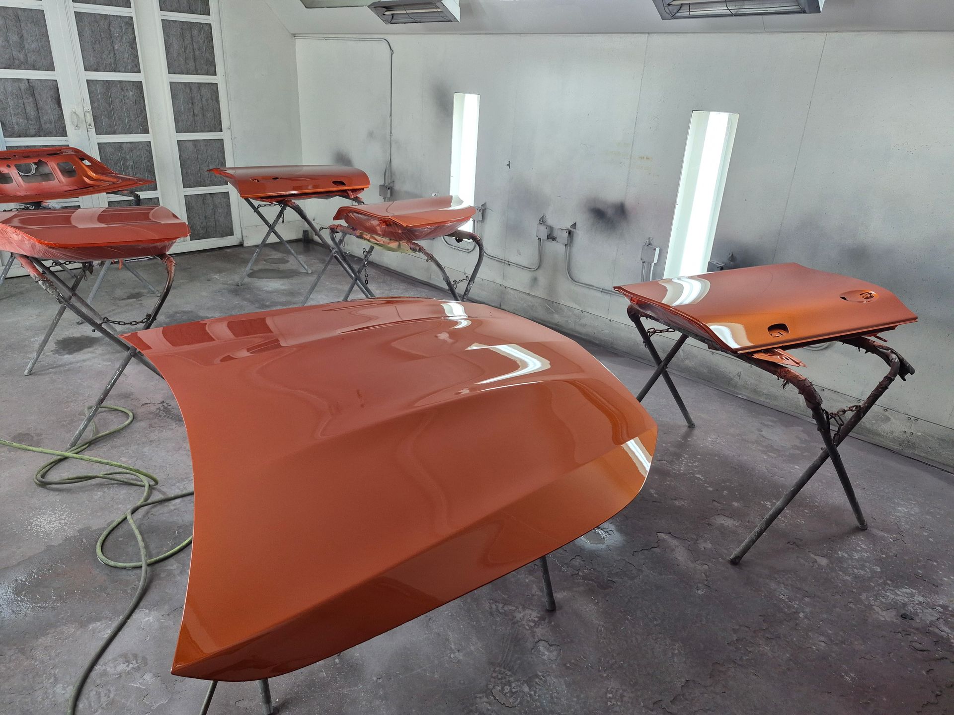 Orange car hood and parts on stands in an auto body shop, with a spray booth in the background
