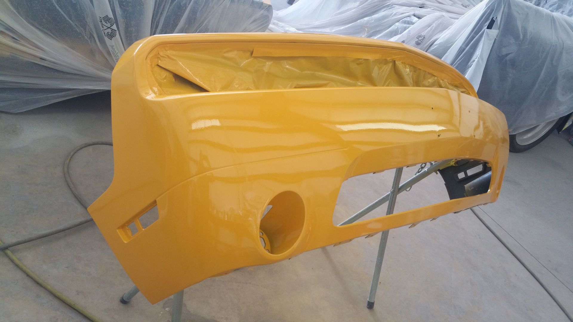 Yellow car bumper hanging in a workshop, with masked parts and paint-spray setup nearby