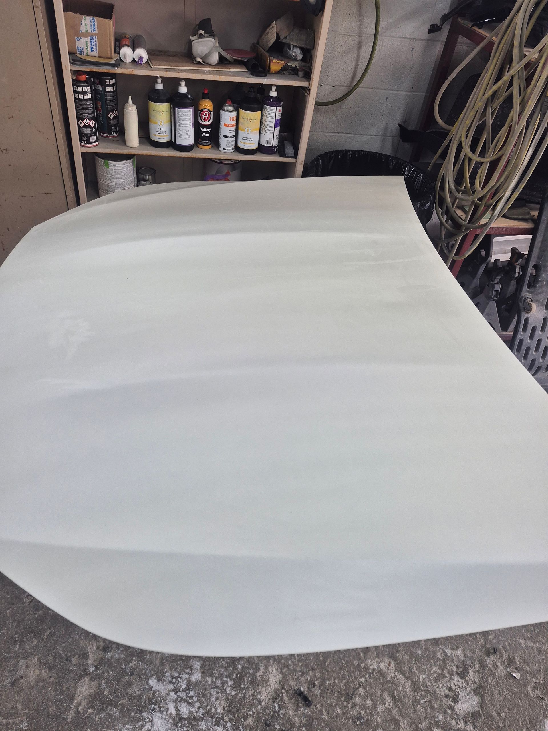 White car hood or body panel in a garage with shelves of supplies in the background