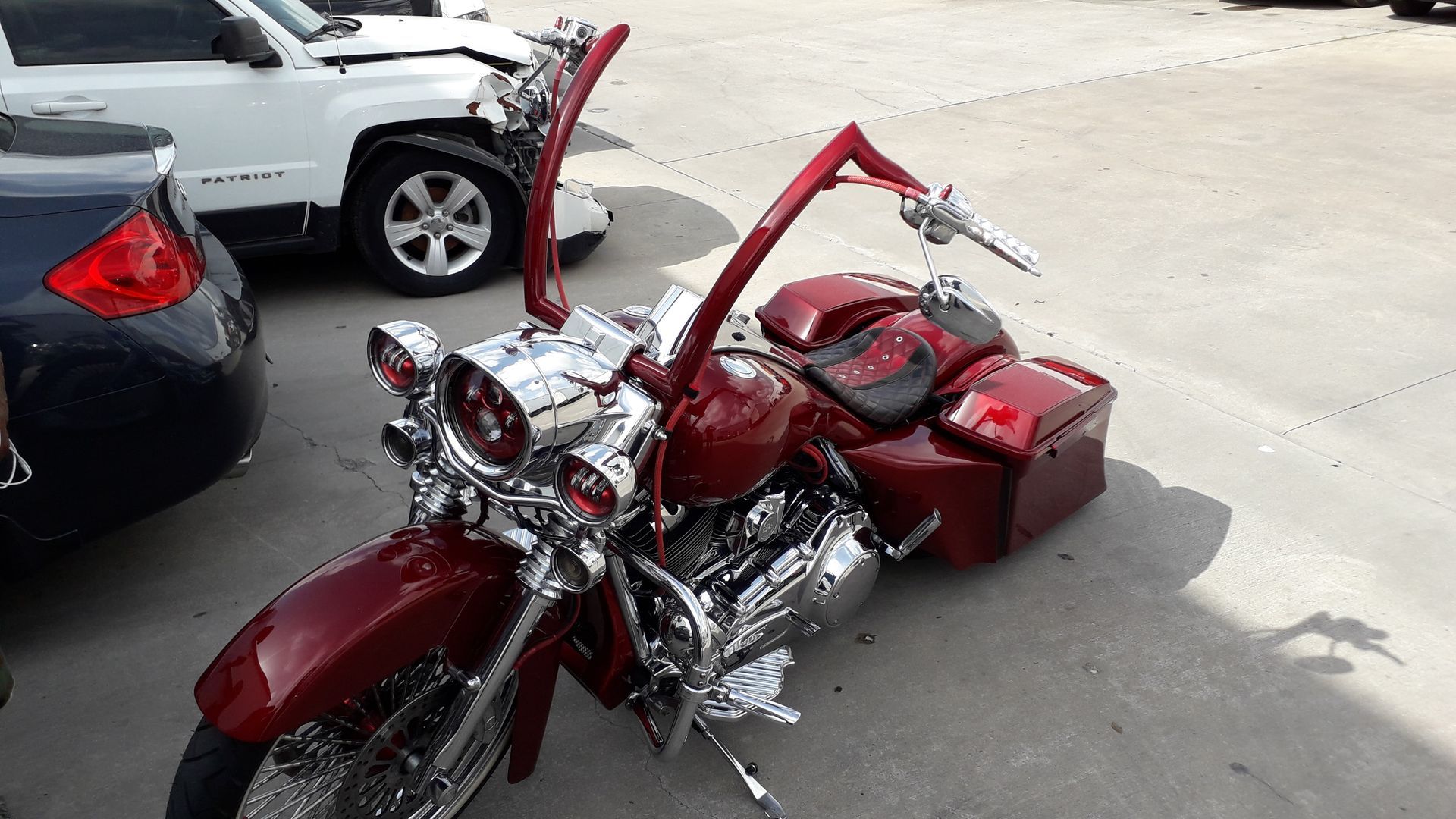 Red custom motorcycle parked beside cars in a parking lot, with tall handlebars and chrome details