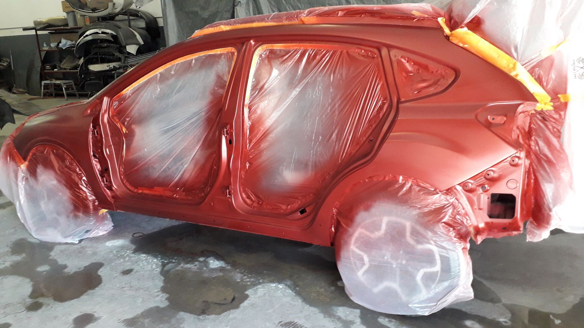 Red car body in a workshop, covered in glossy red paint with masked plastic-wrapped wheels.