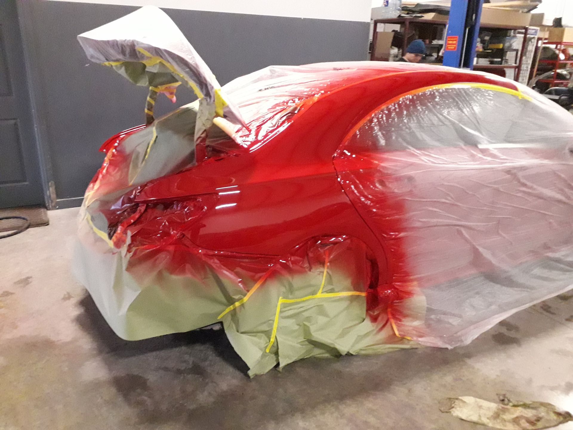 Red car body under repair, partially wrapped in plastic and masking tape in a workshop.