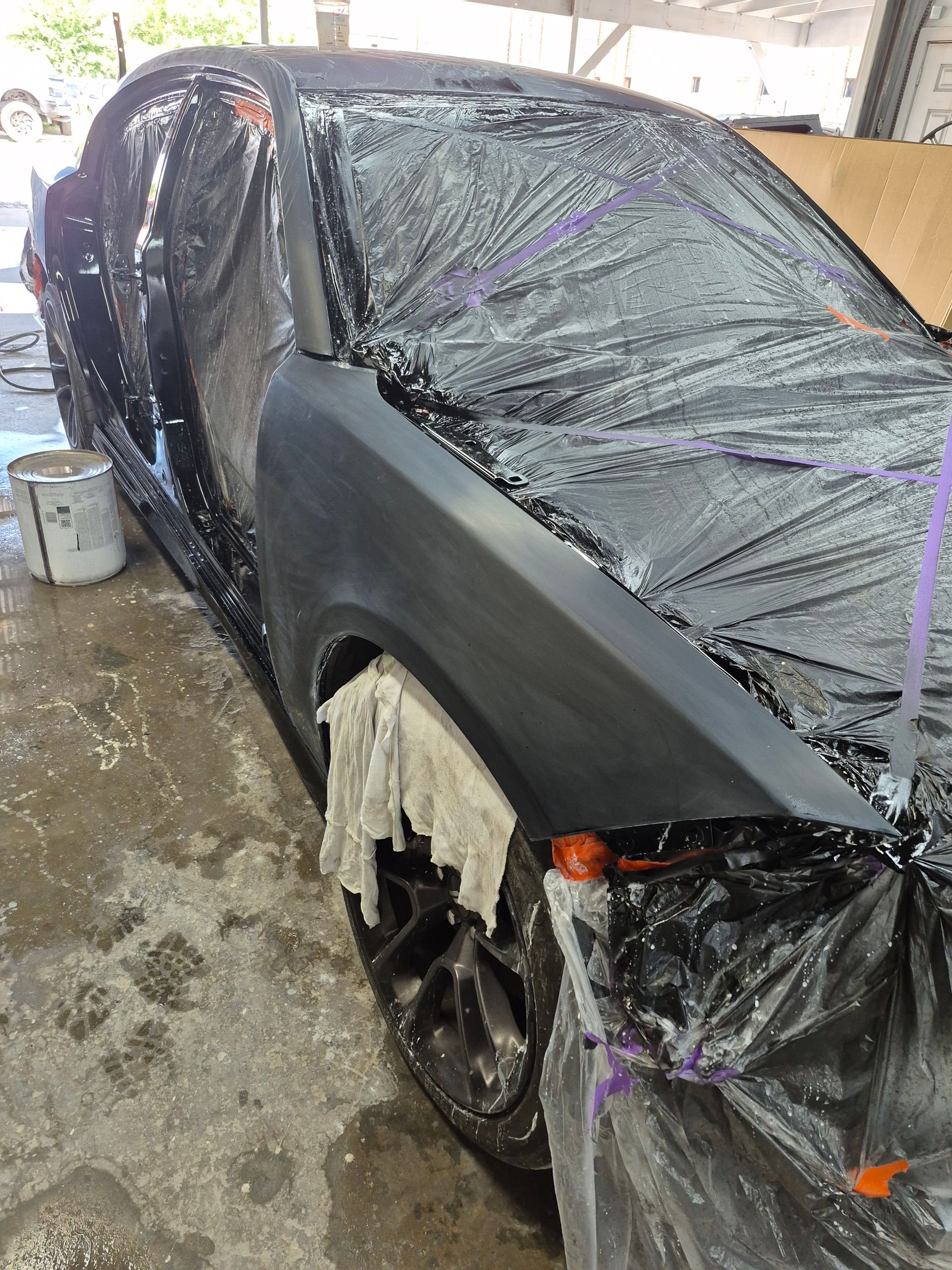 Black car wrapped in plastic and tape, with damaged front wheel in a wet garage.