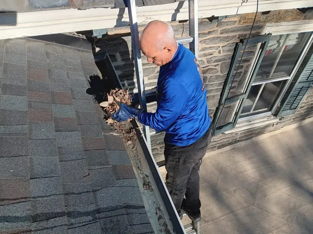 Worker in blue shirt cleaning a gutter on a roof beside a brick house