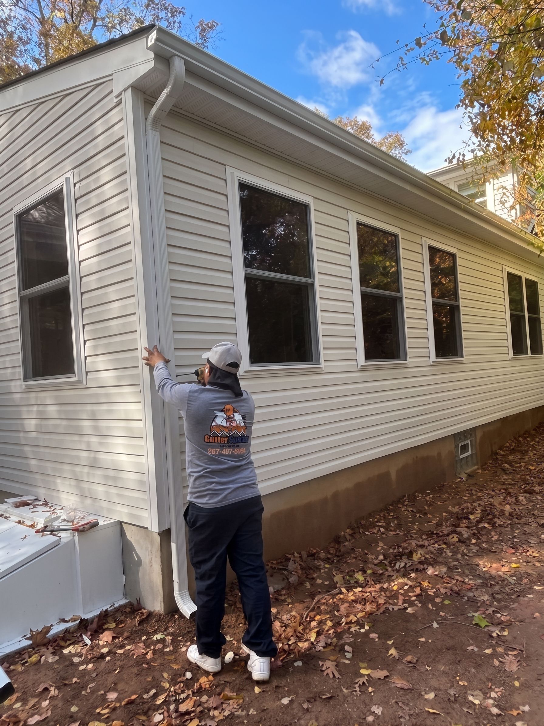 Person inspecting white house siding with a window under repair in a leaf-covered yard