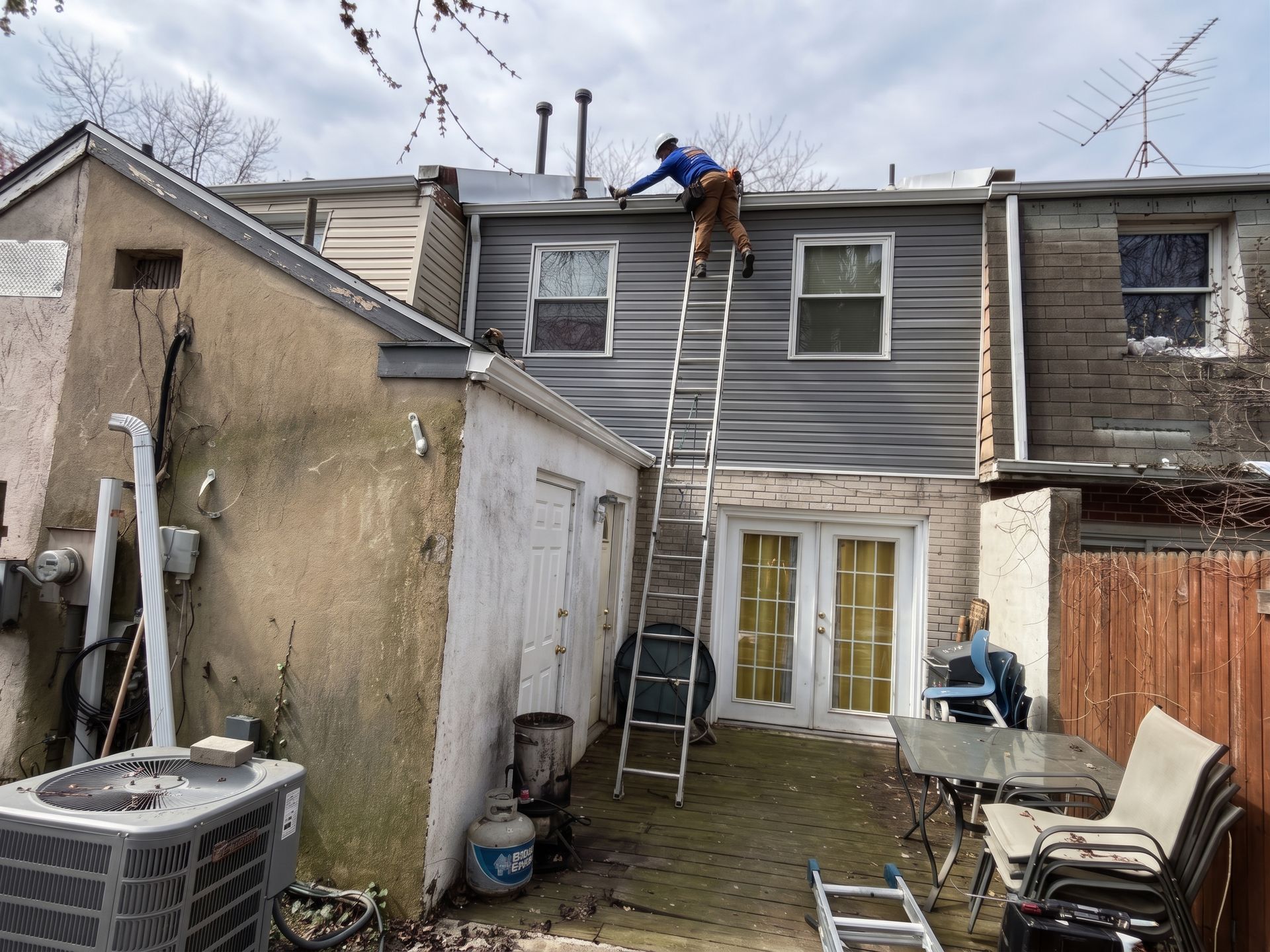 Worker on ladder repairing the roof of a gray row house in a narrow backyard.
