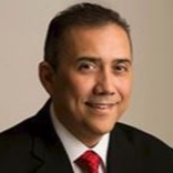Studio headshot of a smiling man in a dark suit, white shirt, and red tie against a beige background