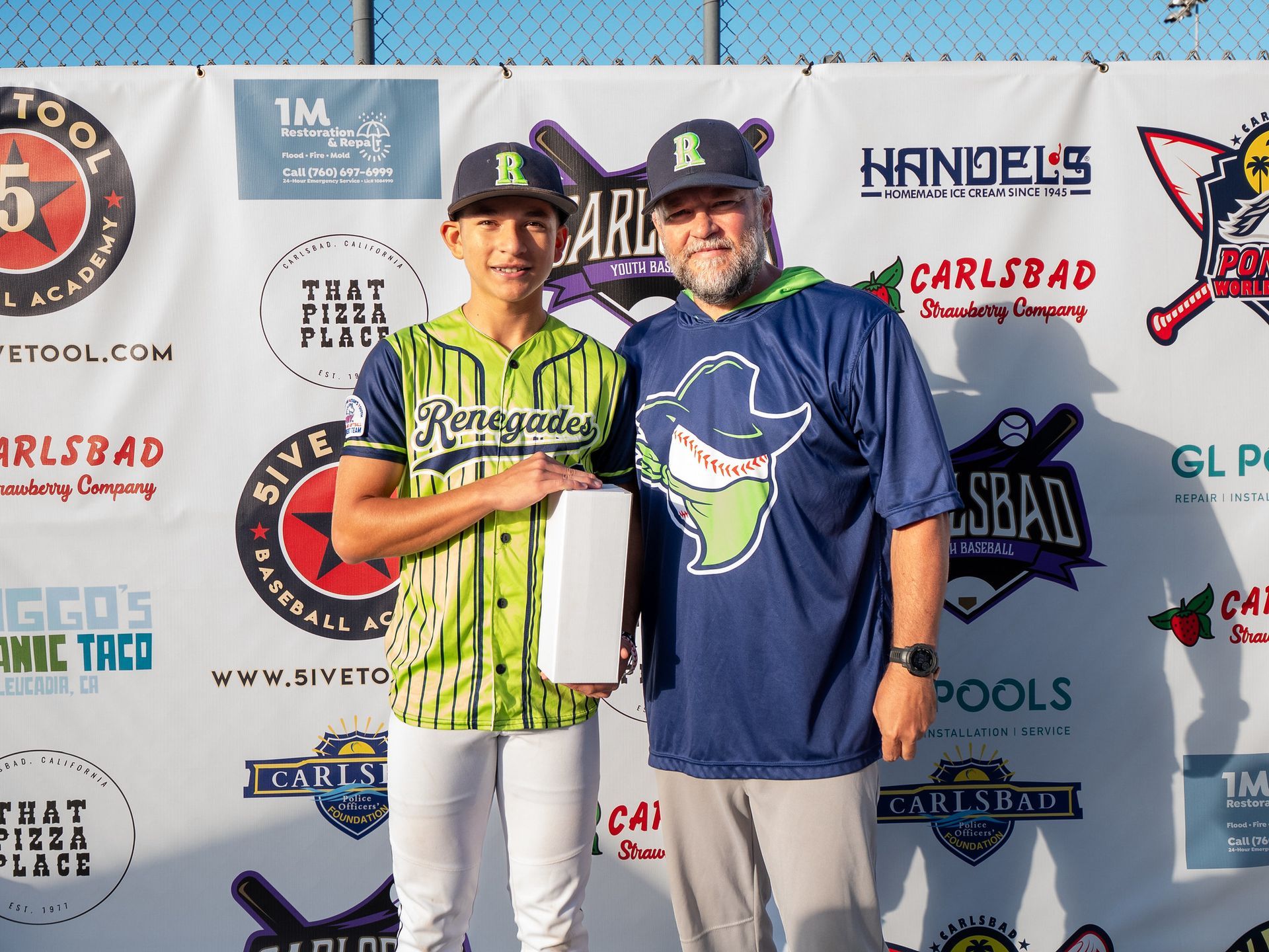 Two men pose holding a trophy at a baseball event backdrop, one in green jersey and one in blue shirt.