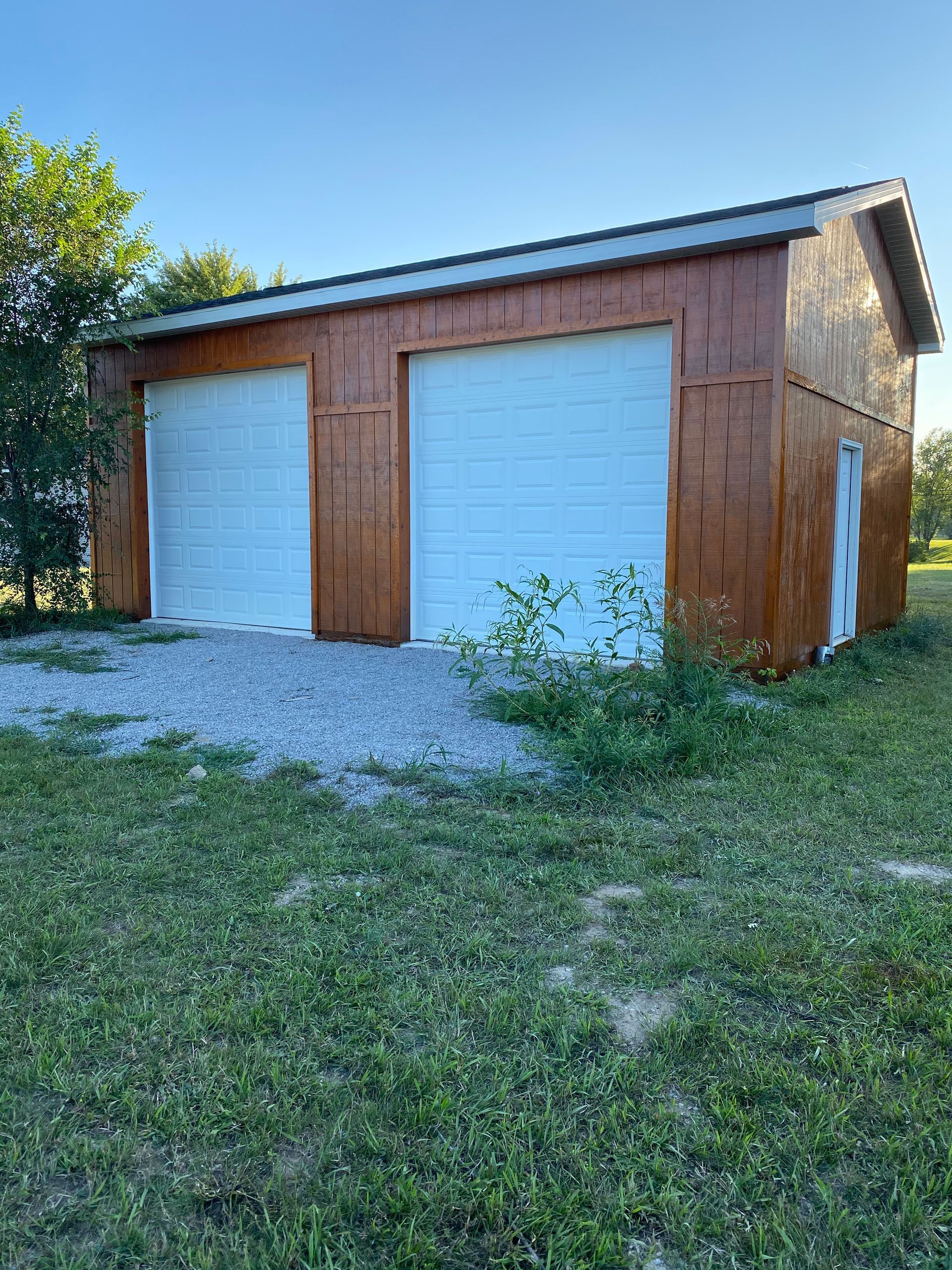 A garage with two white garage doors is sitting in the middle of a grassy field.