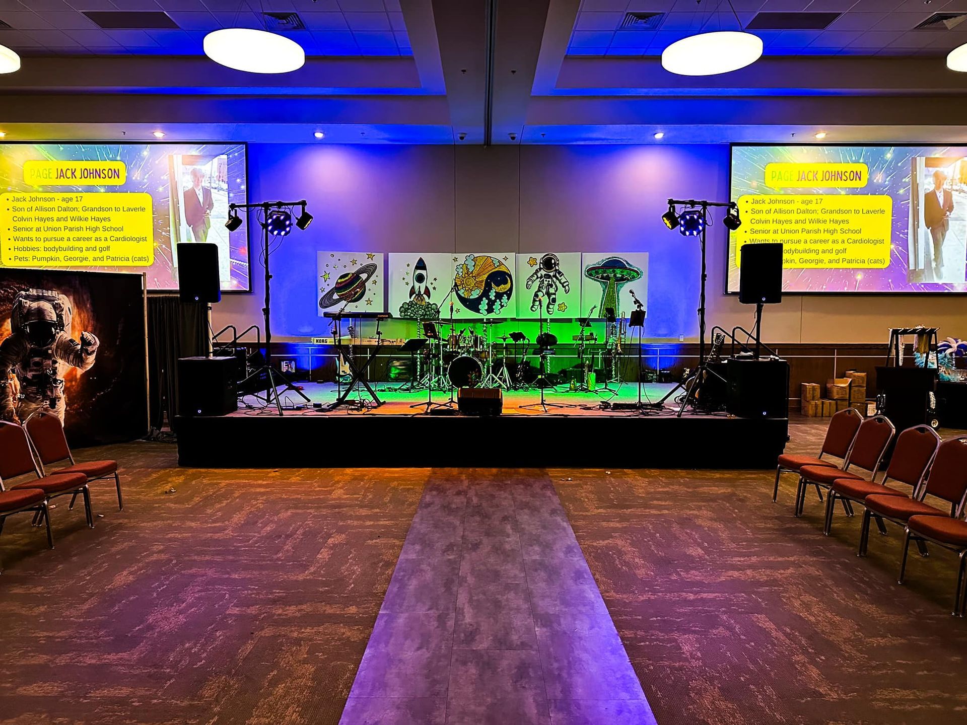 A stage in a conference room featuring musical equipment, artwork, and side screens, with seating rows on either side.
