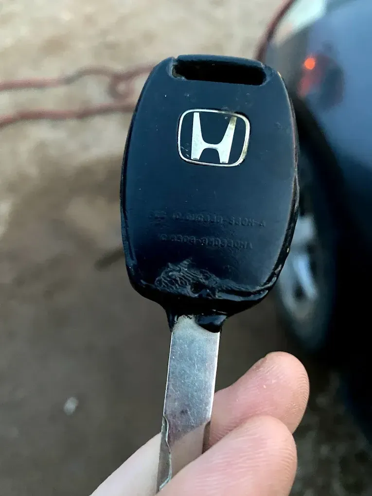 Black Honda car key with the logo held in a hand near a car.