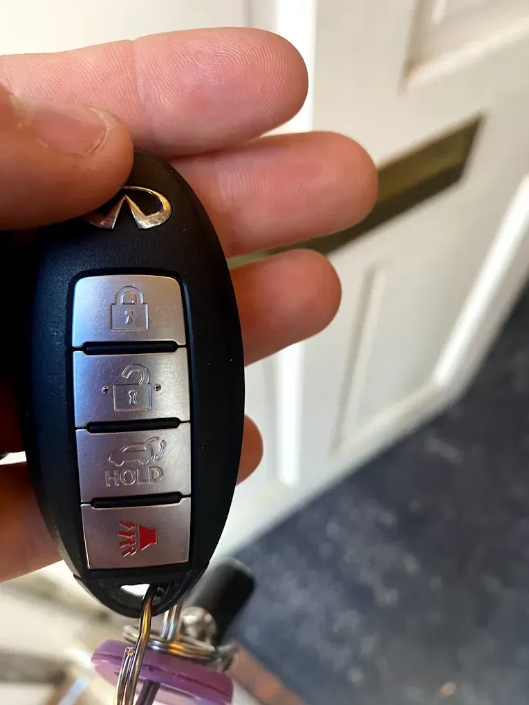 Hand holding an Infiniti car key fob, with lock, unlock, panic, and car start buttons.