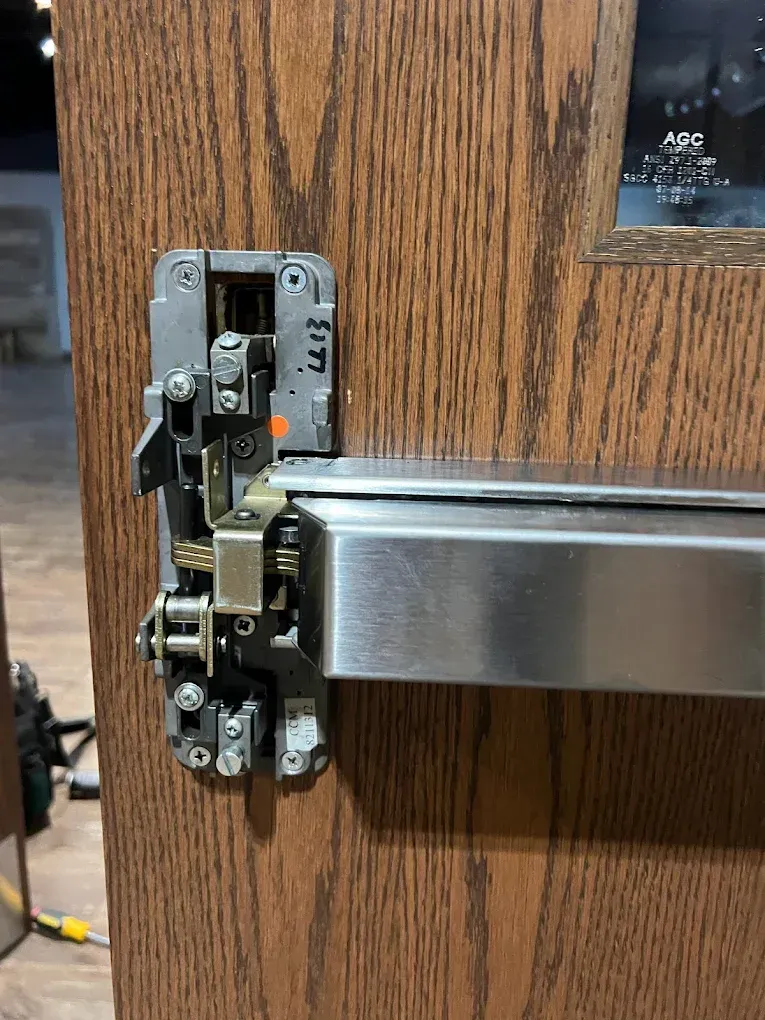 Metal push bar door hardware on a wood door, partially disassembled.
