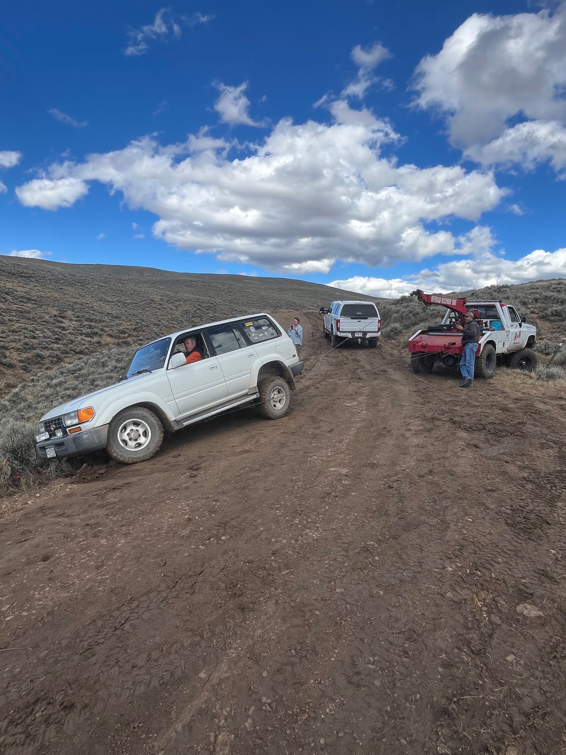 White SUV on a dirt road, others nearby. Blue sky with clouds.