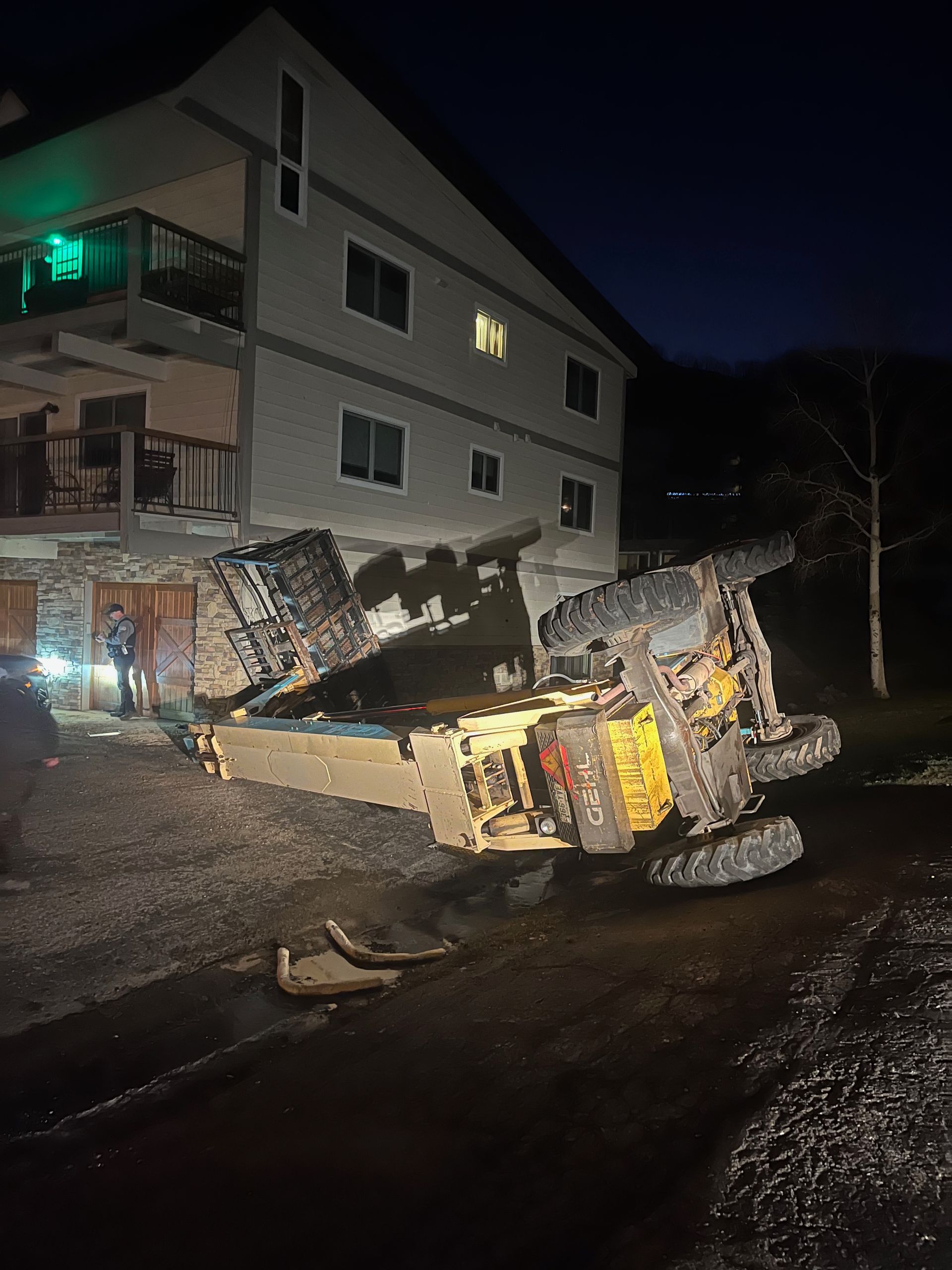 A construction vehicle overturned near a house at night; person stands near vehicle.