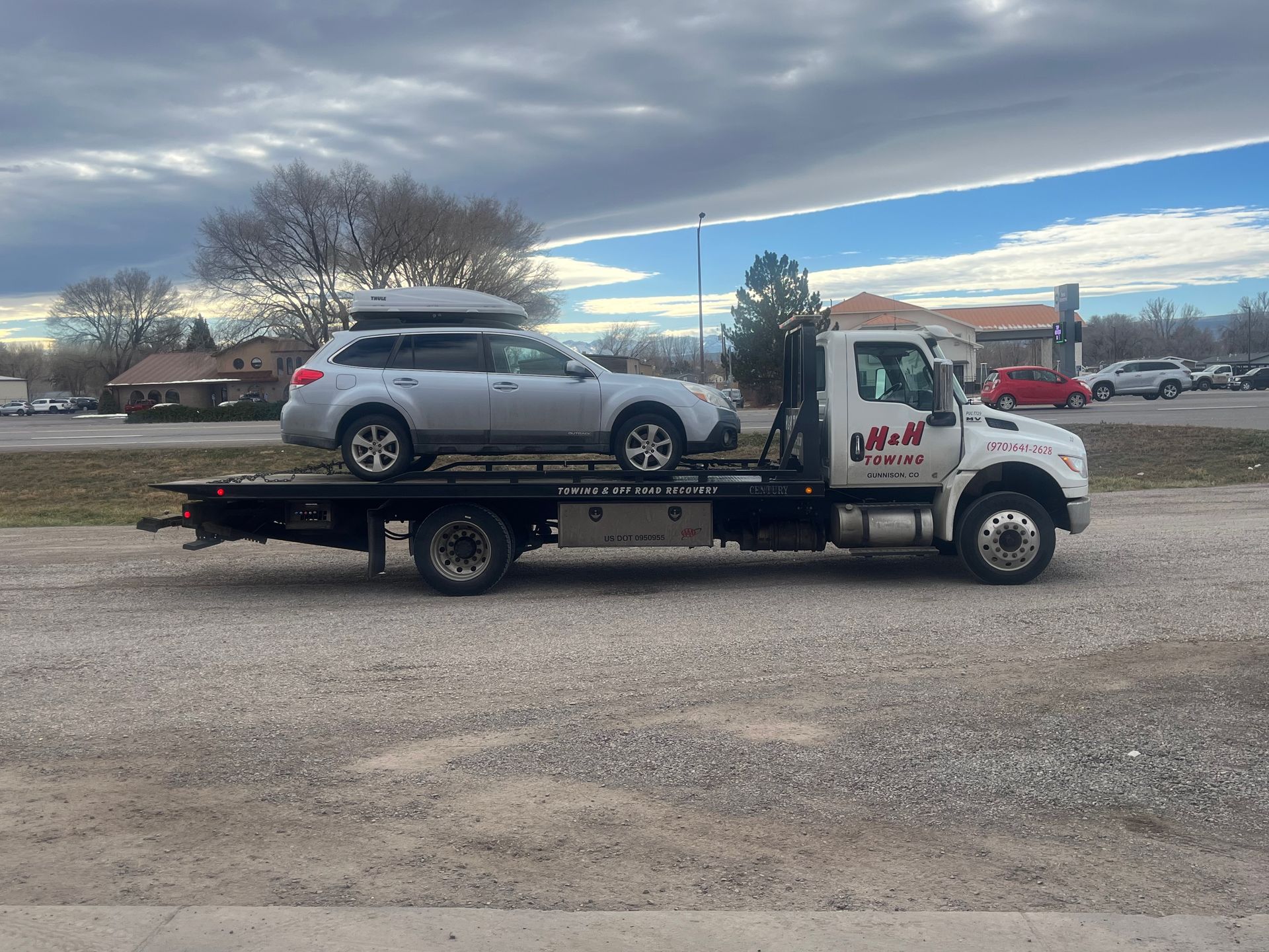 Silver Subaru Outback on a flatbed tow truck. White truck with black writing parked outside. Cloudy sky.