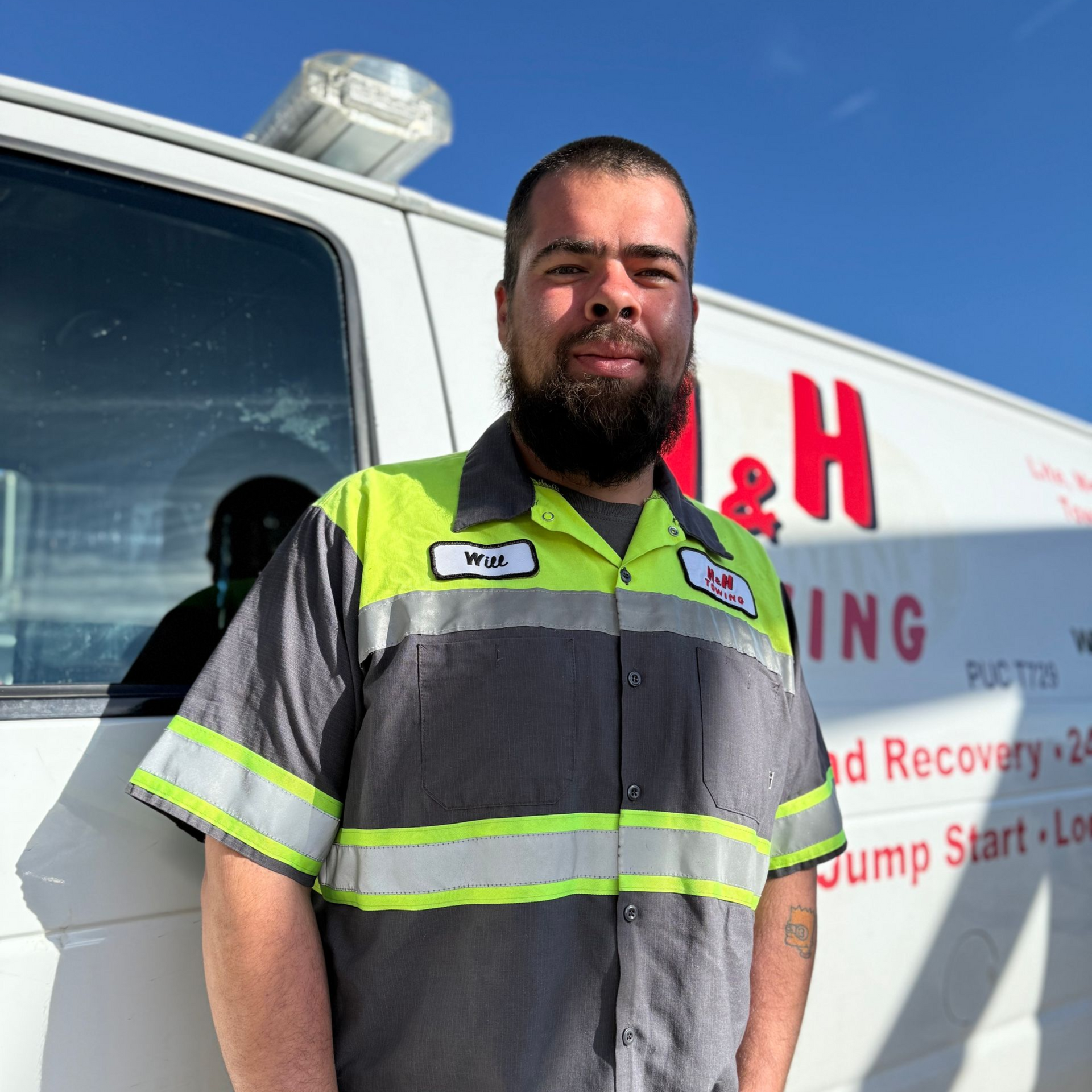 Man in work uniform stands near a tow truck. The truck has