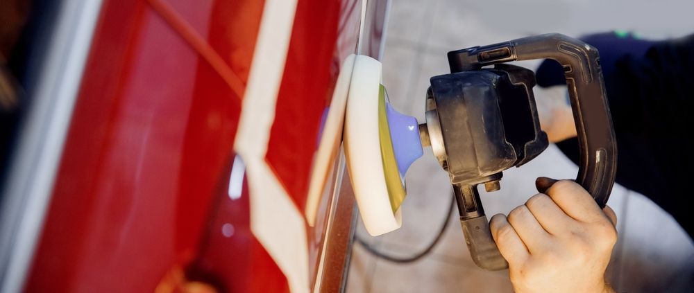 A hand holding an electric buffer tool against a red car panel to polish the surface.