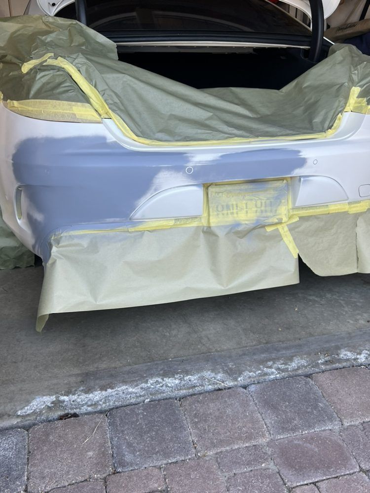 The rear bumper of a white car prepared for painting, with grey primer applied and surrounding areas masked with tape.