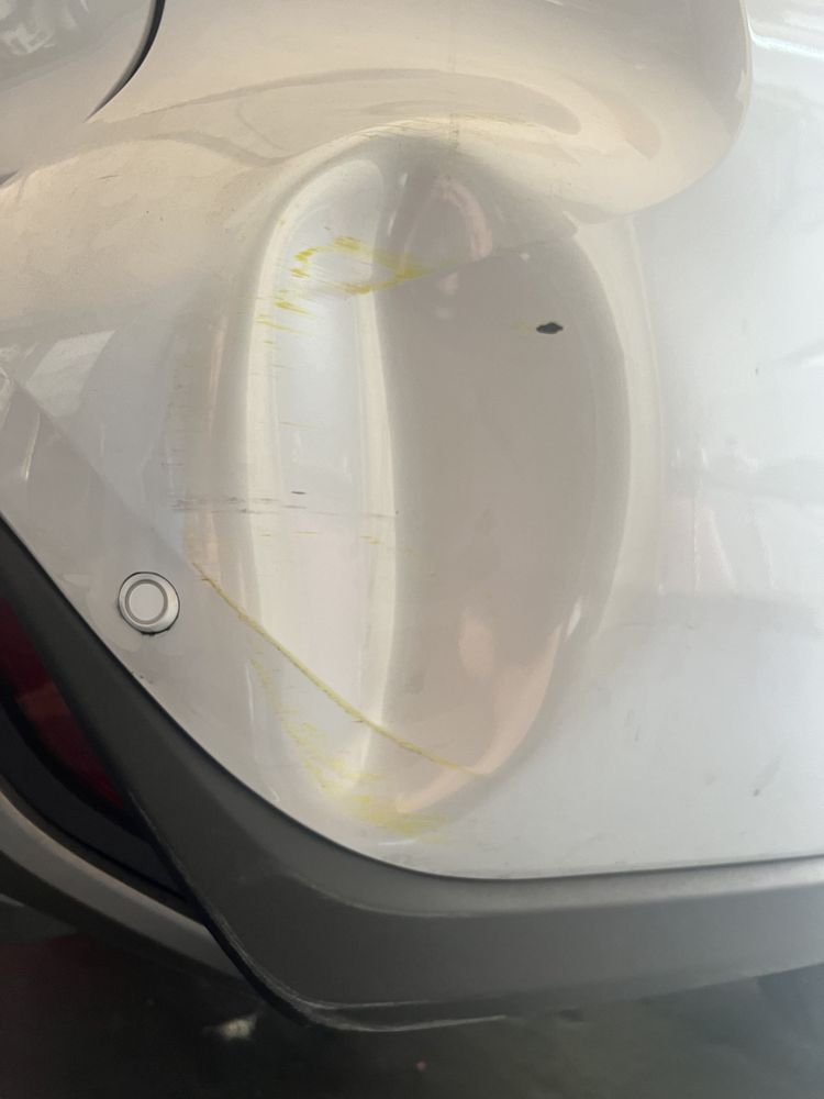 A close-up of a white car bumper showing a dent and yellow paint transfer marks near a parking sensor.