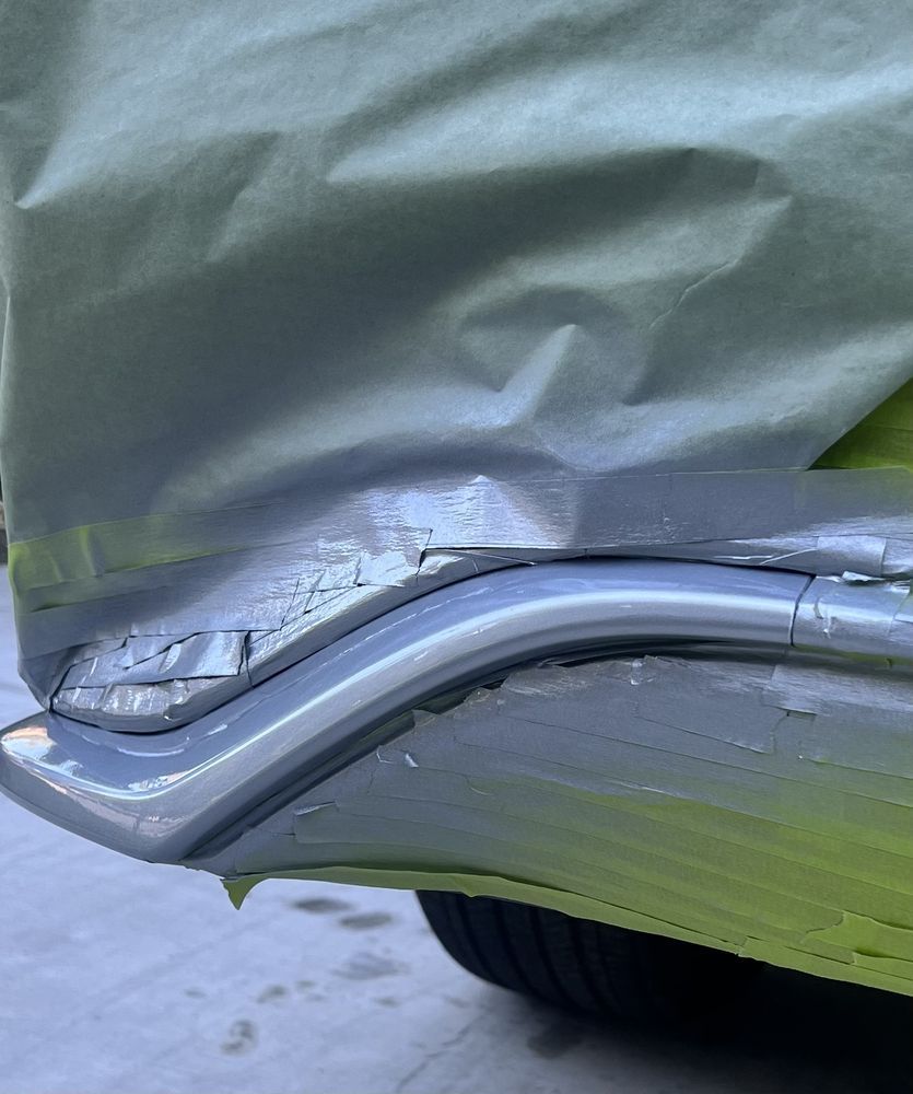 A close-up view of a car's side panel with grey paint being applied, partially masked with green tape and paper.