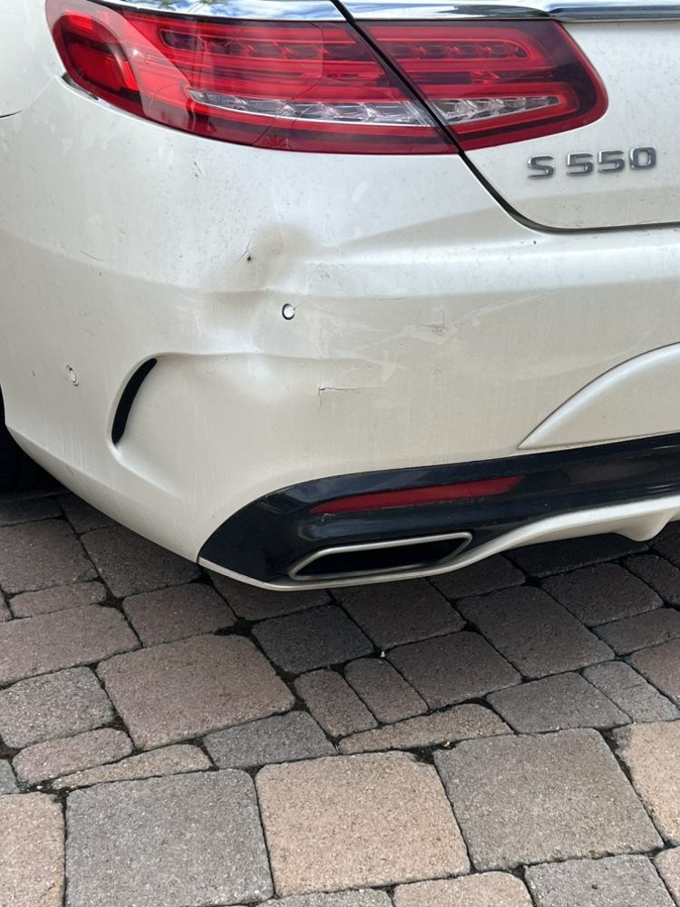A white Mercedes S 550 with a dented rear bumper parked on a stone paver driveway.