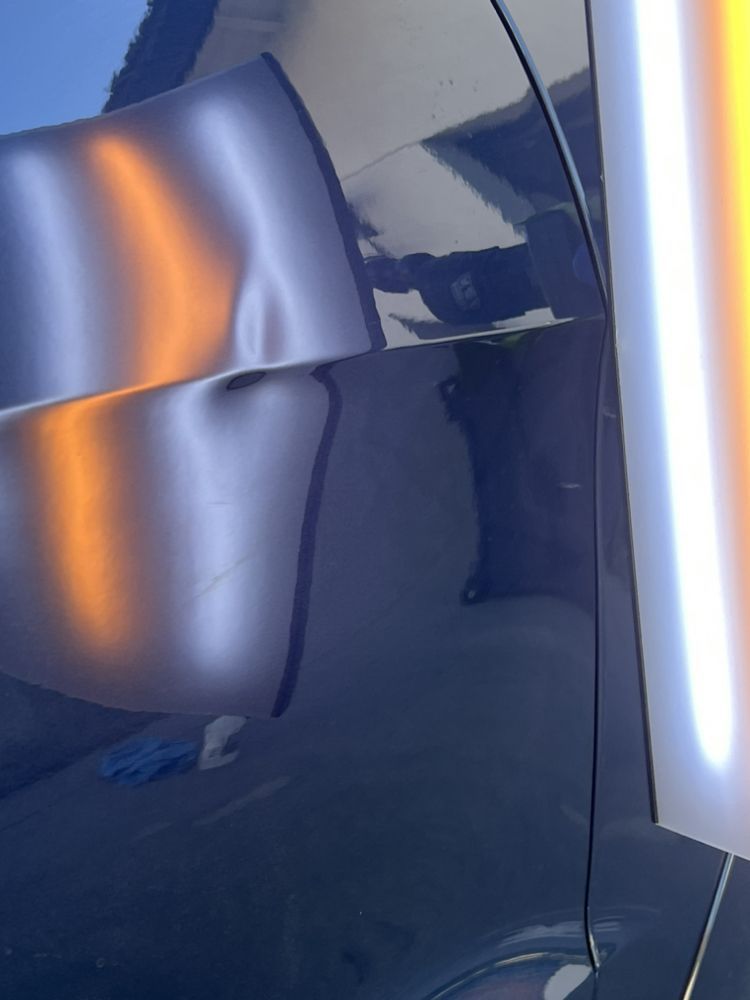A dark car panel showing a dent highlighted by the reflection of a bright, linear shop light used for repair assessment.
