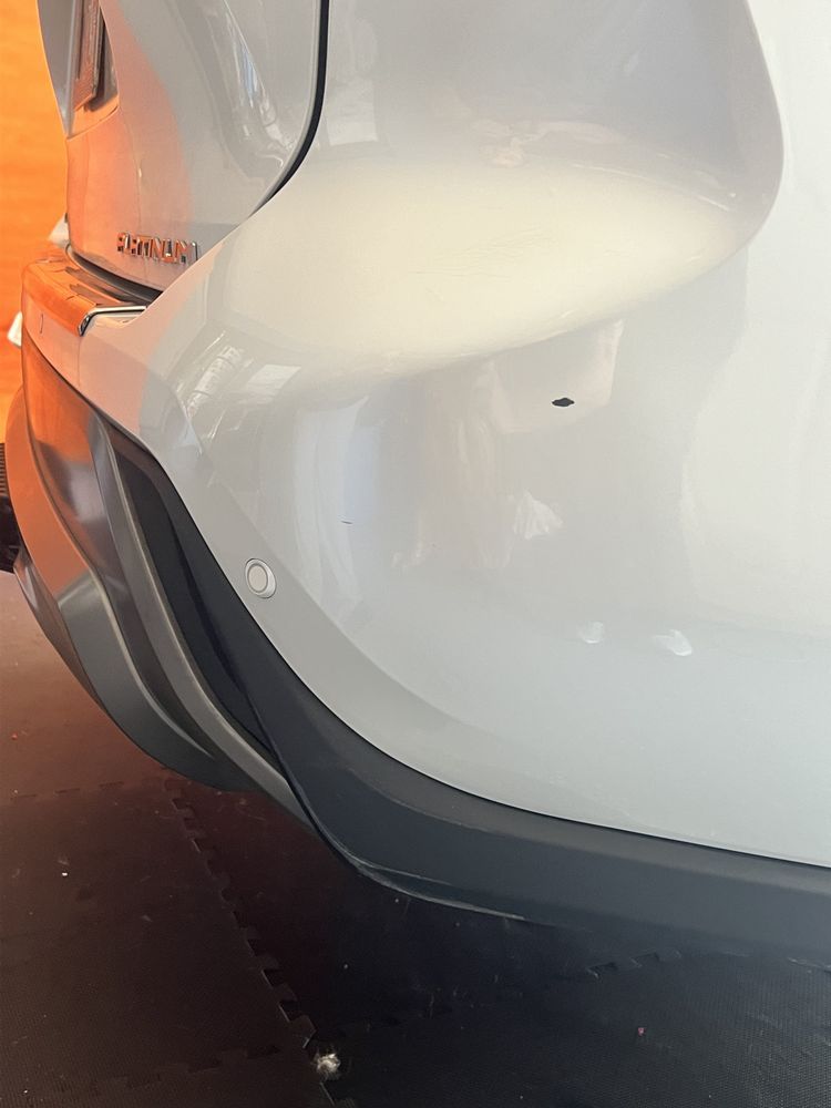 Close-up of a white car's rear bumper showing a parking sensor and a small dark mark on the paint.