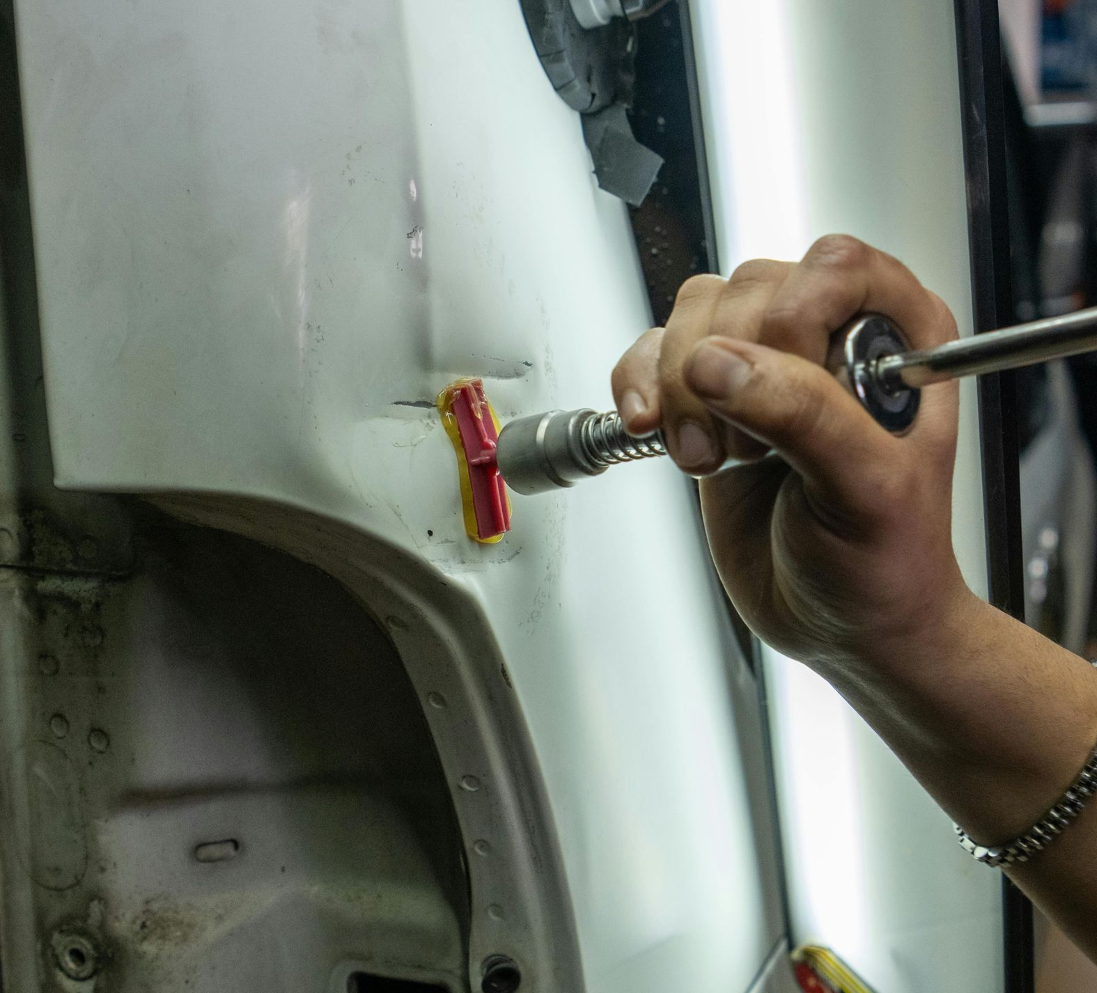 A hand uses a specialized tool to perform paintless dent repair on a white car door panel.