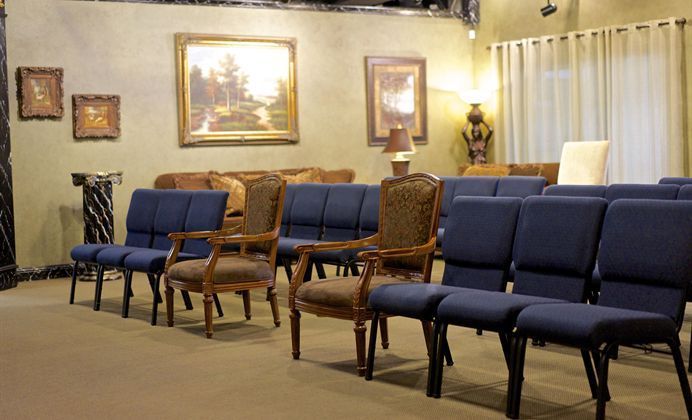 Empty waiting room with rows of blue and wooden chairs, framed wall art, and curtains.