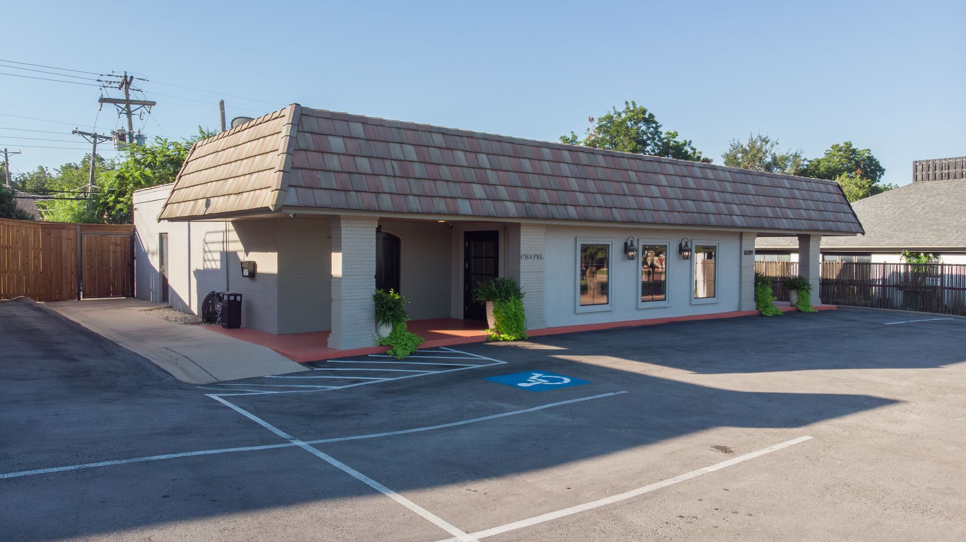 Single-story beige building with a shingled roof and windows, seen across an empty parking lot.