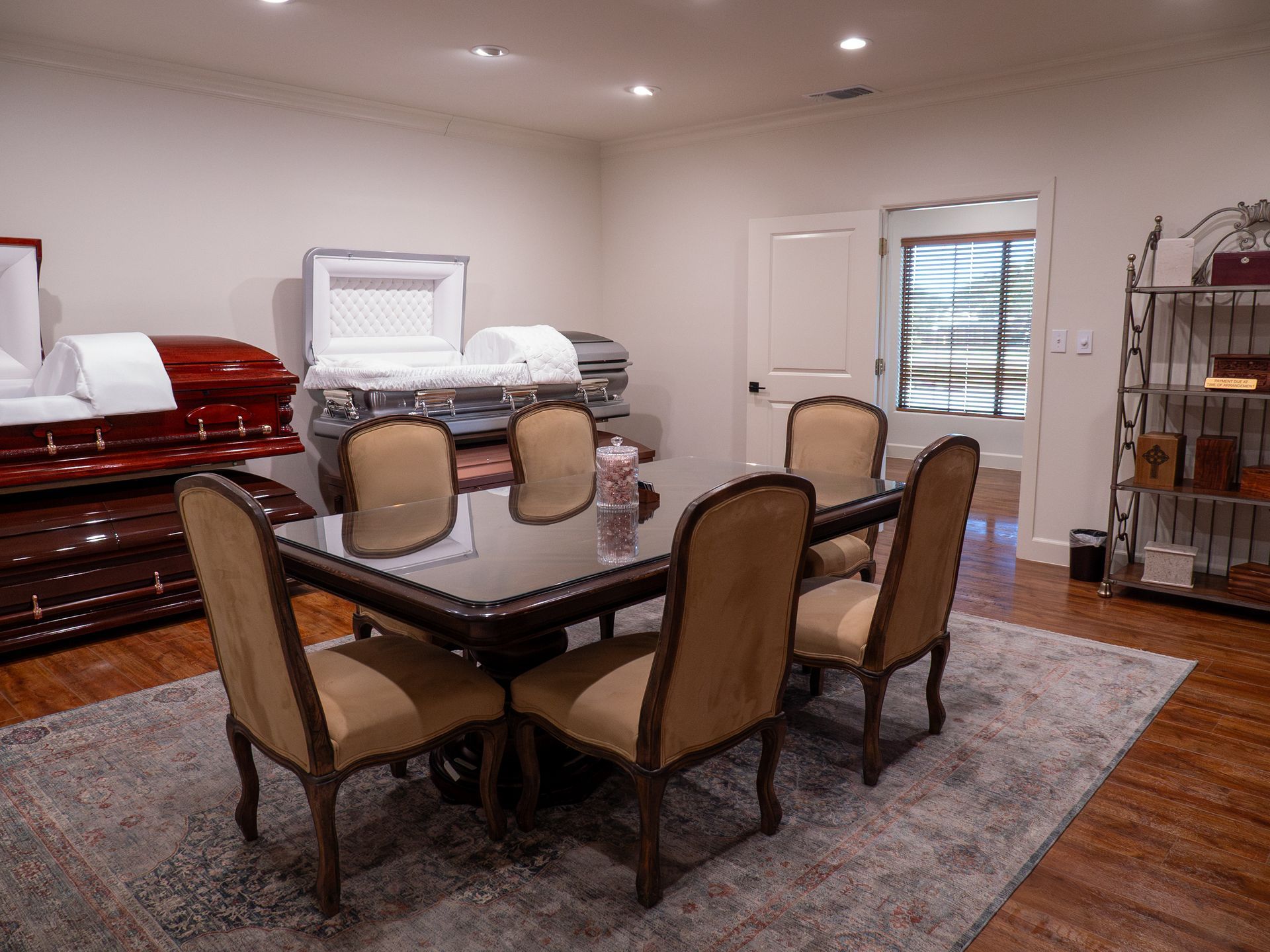 Casket showroom with two open caskets, dining table, chairs, and display shelves on a carpeted floor