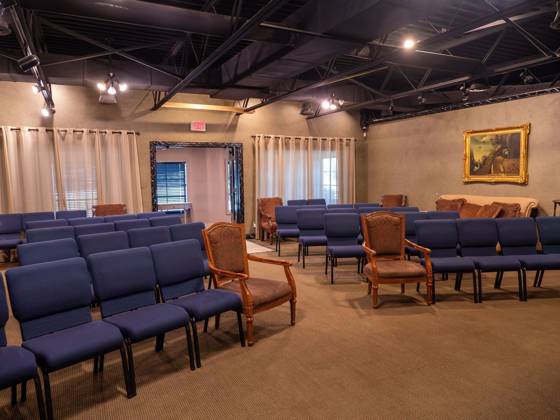 Empty waiting room with rows of blue chairs, beige walls, curtains, and a framed painting.