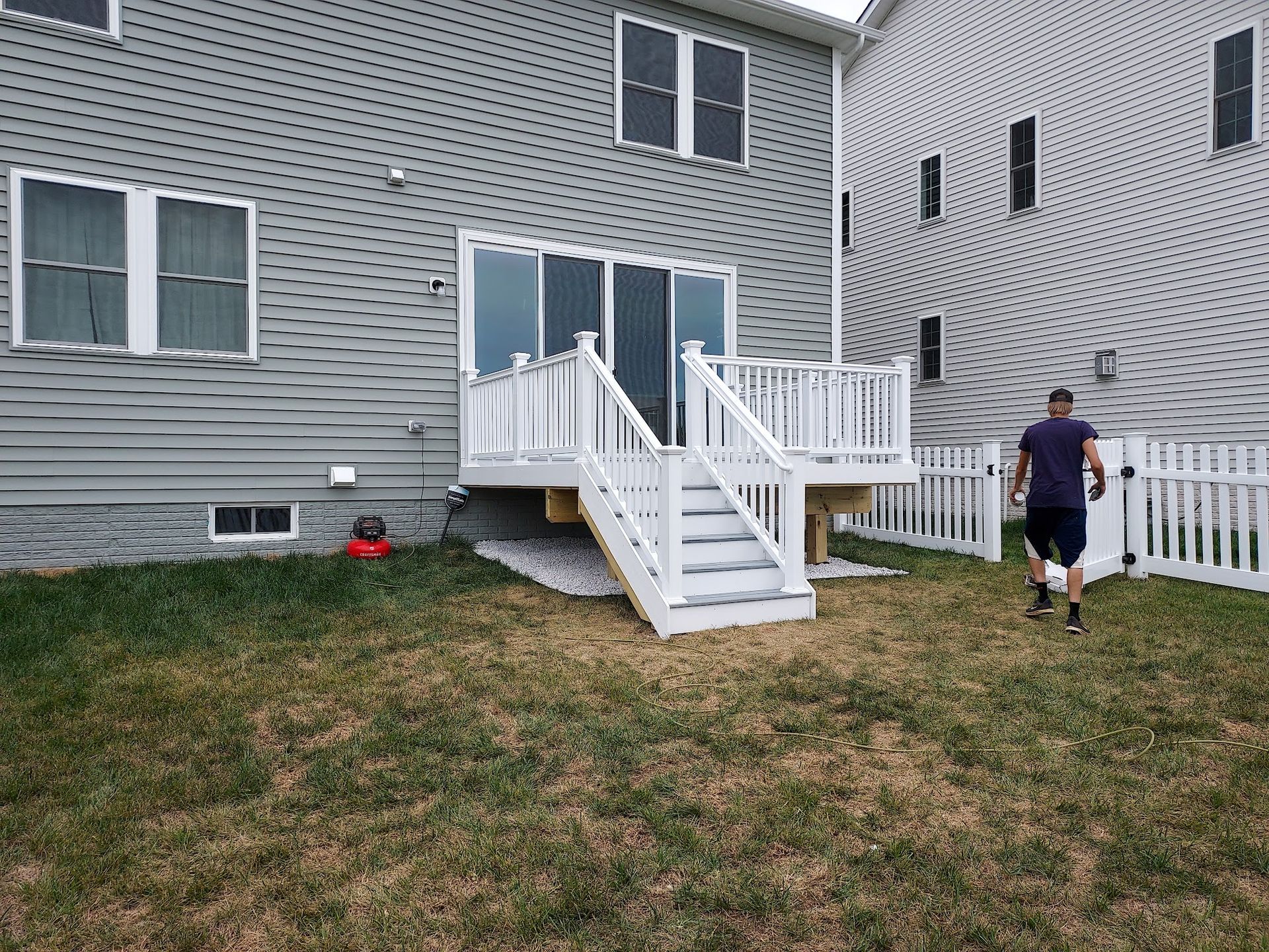 Backyard with a white deck and fence. A person walks on the grass toward the right. Grey houses surround.