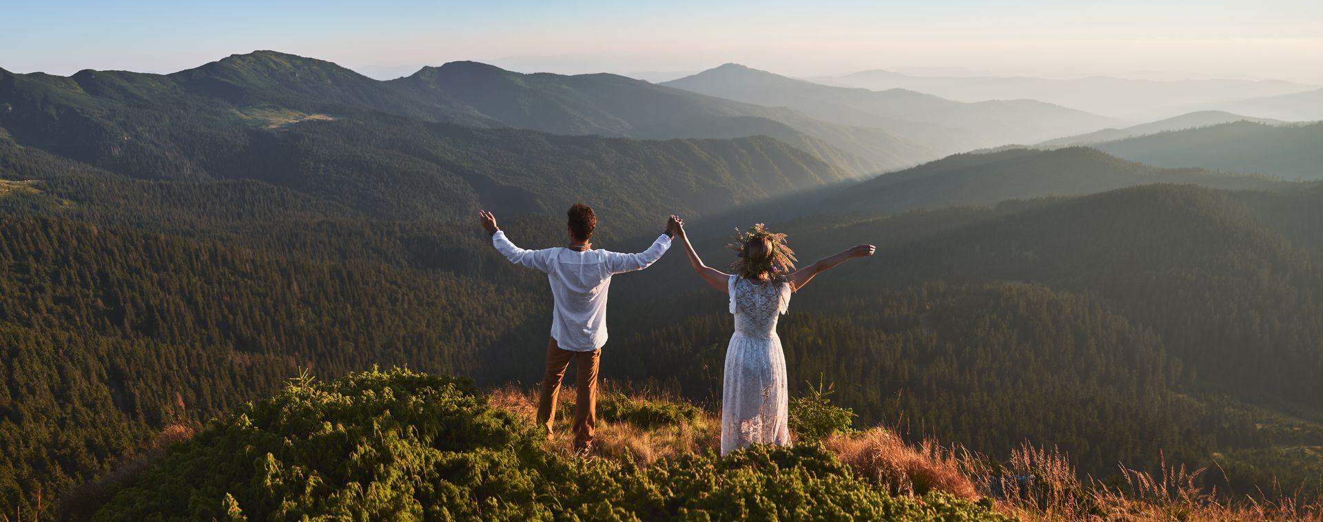 Two hikers stand on a mountain overlook with arms outstretched, facing a sunlit forested valley.