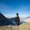 Person sitting on a rocky ledge overlooking mountains and clouds under a blue sky