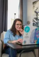 Woman on a video call, waving at a laptop in a bright room with curtains and a potted plant.
