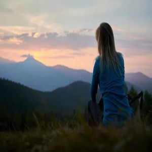 Person sitting in a field at sunset, looking toward distant mountains and a pink sky