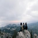 Three hikers sit on a rocky peak overlooking a cloudy mountain valley and distant coastline