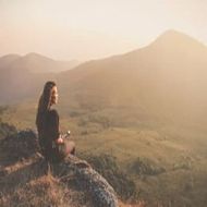 Person sitting on a rocky overlook, gazing over a sunlit mountain valley at sunset