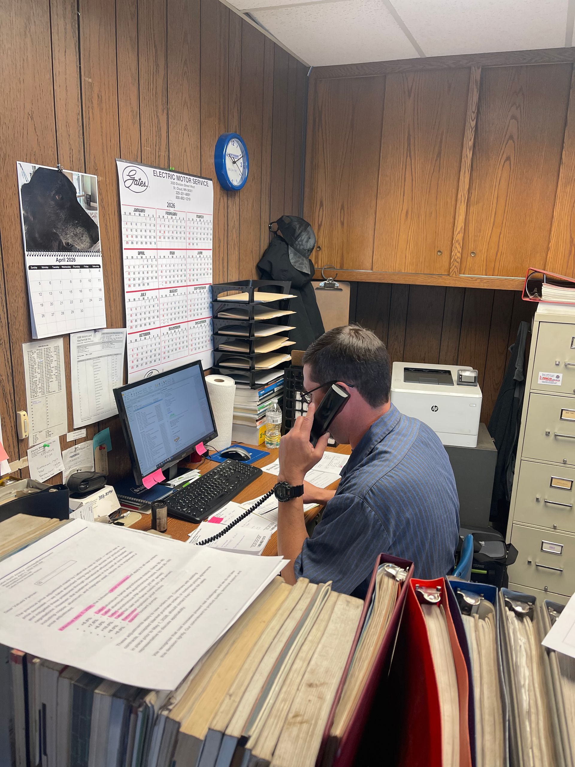 A person in a blue shirt sits at a cluttered desk in a wood-paneled office, talking on a phone while looking at a monitor. A person in a blue shirt sits at a cluttered desk in a wood-paneled office, talking on a phone while looking at a monitor.