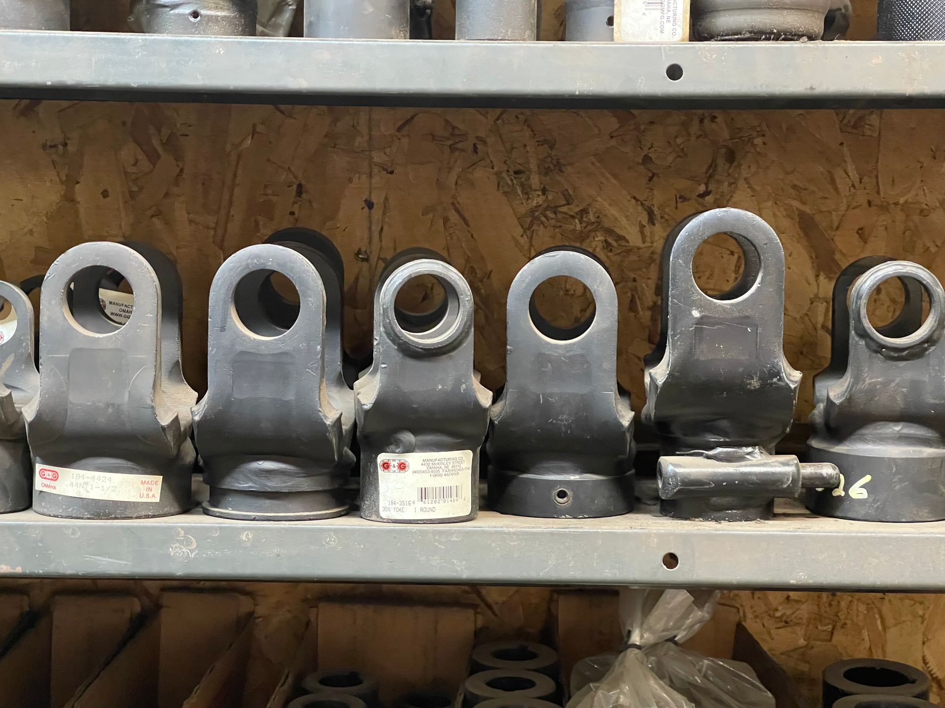 A row of dark metal clevis ends sits on a storage shelf in a shop, with some pieces bearing small white inventory labels.